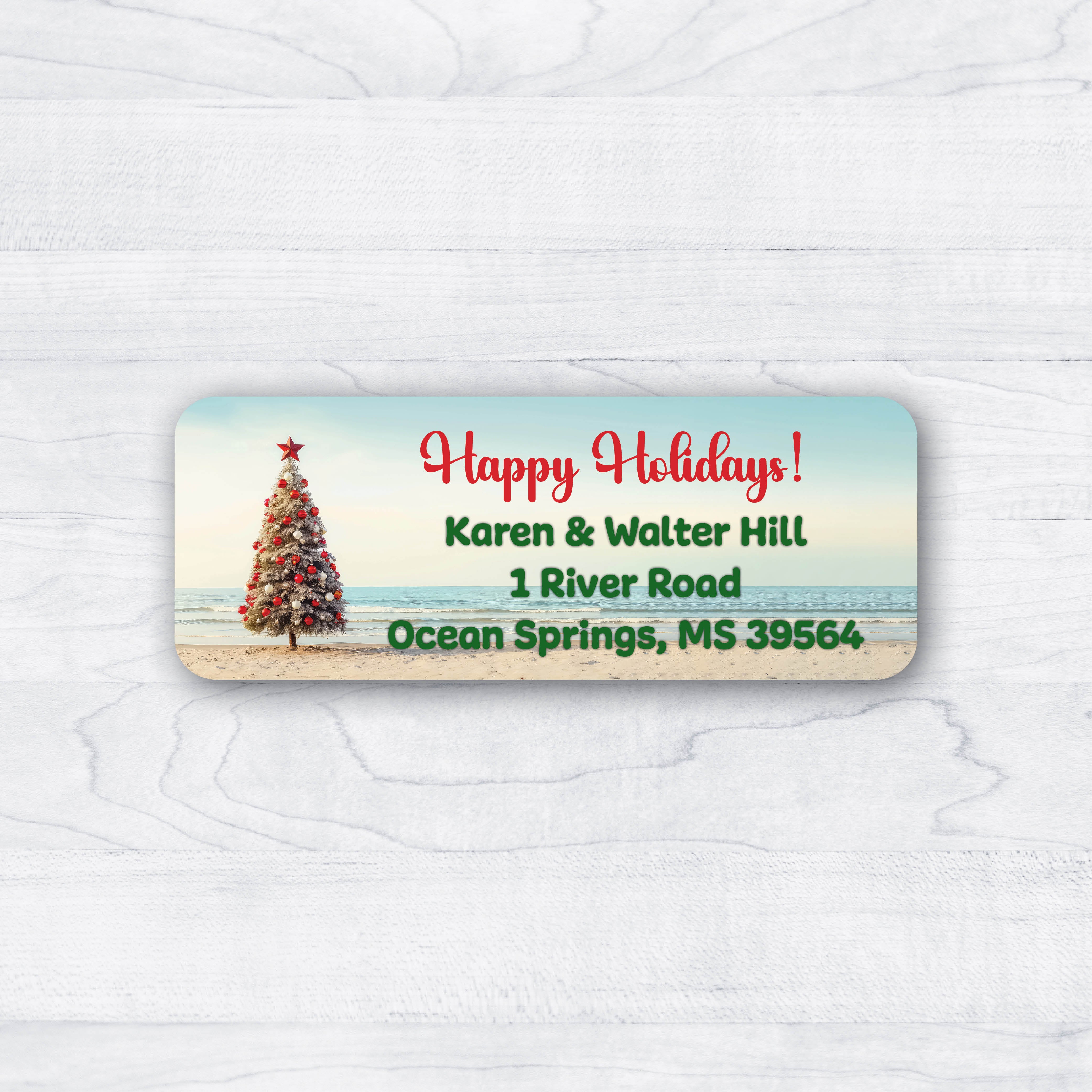 A rectangular holiday address label with rounded corners, featuring a beach scene with a decorated Christmas tree, blue sky, and ocean. The label includes the text 'Happy Holidays!' followed by a name and address in red and green lettering, shown on a white wood-textured background.