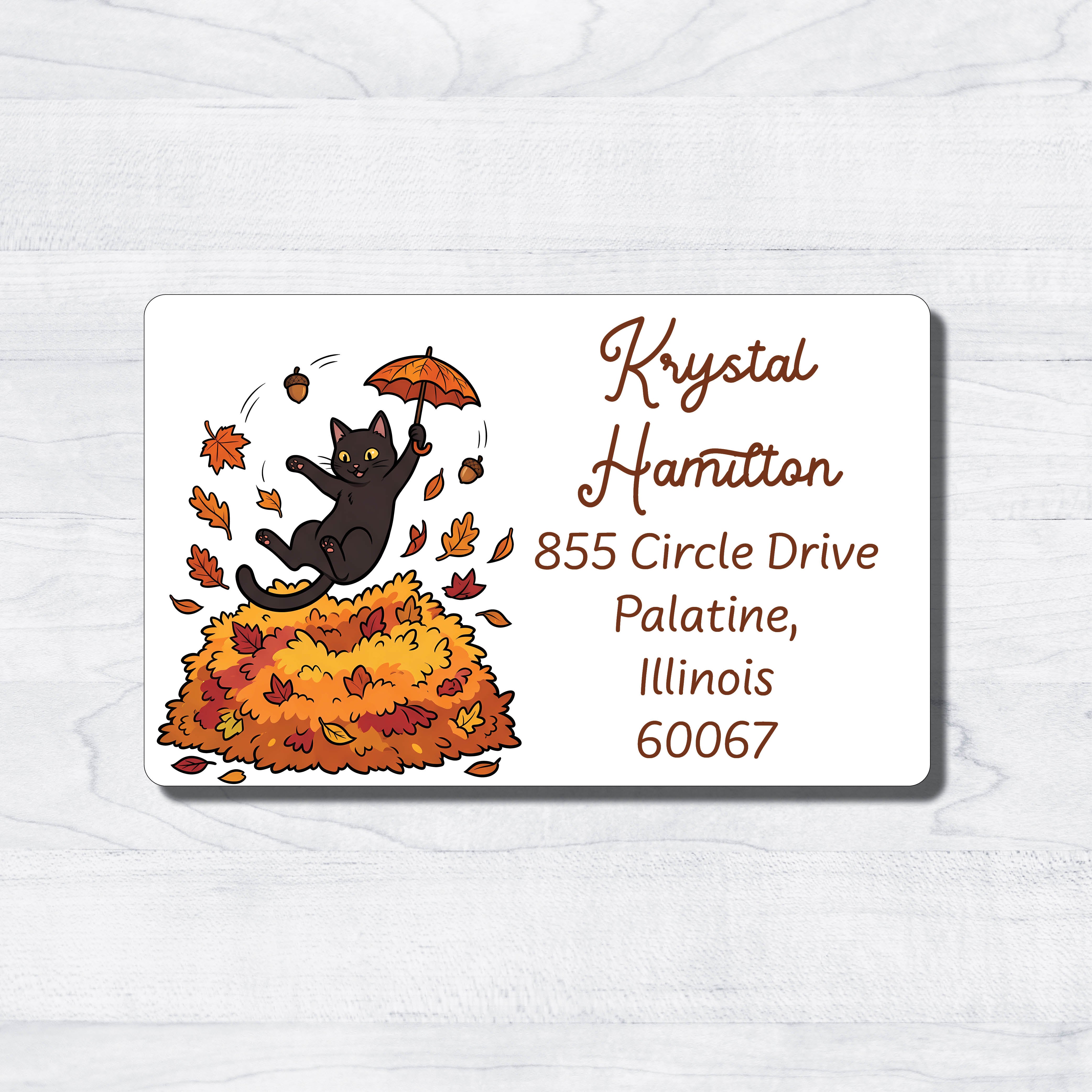 A rectangular address label with rounded corners, featuring a black cat jumping on a pile of orange and yellow autumn leaves while reaching for a small orange umbrella. The right side of the label has sample address text in brown on a white background, placed on a light wood surface.