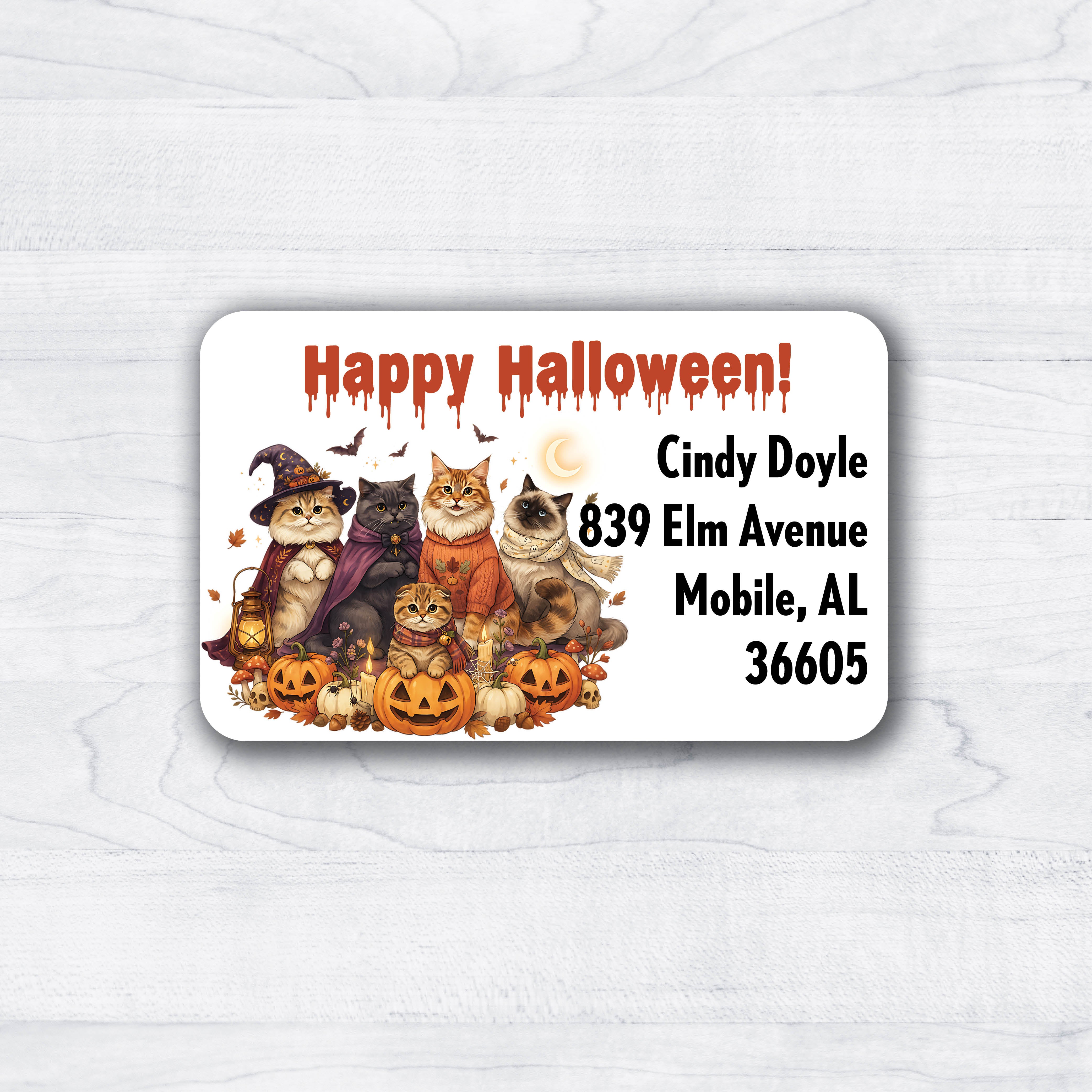 A rectangular Halloween-themed address label with rounded corners, featuring illustrated cats dressed as witches and pumpkins, the text 'Happy Halloween!' in dripping orange letters, and sample address text on a white wood background.
