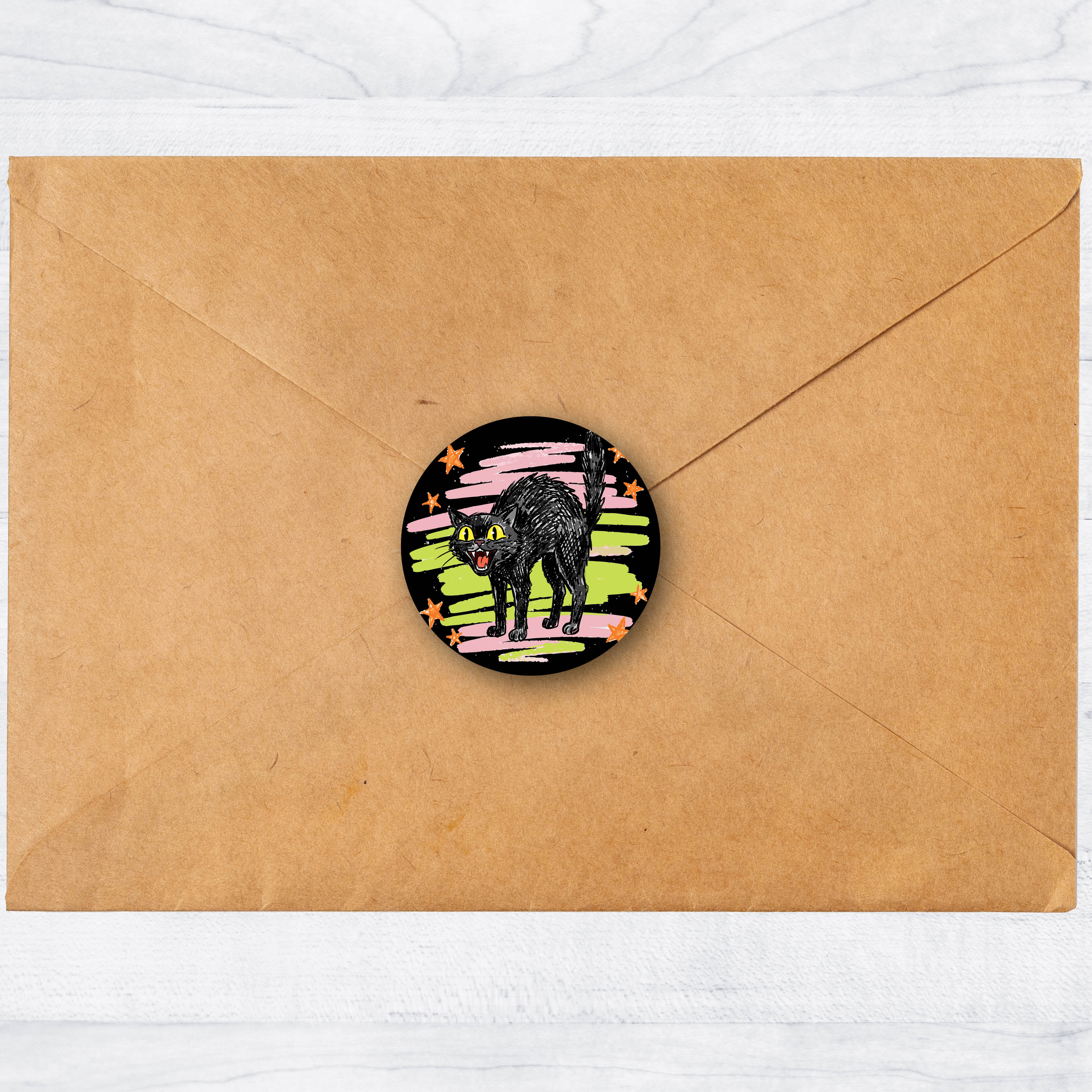 A round sticker featuring a hissing cat illustration on a colorful pink, green, and black background, shown on top of a brown kraft paper envelope against a white marble surface.