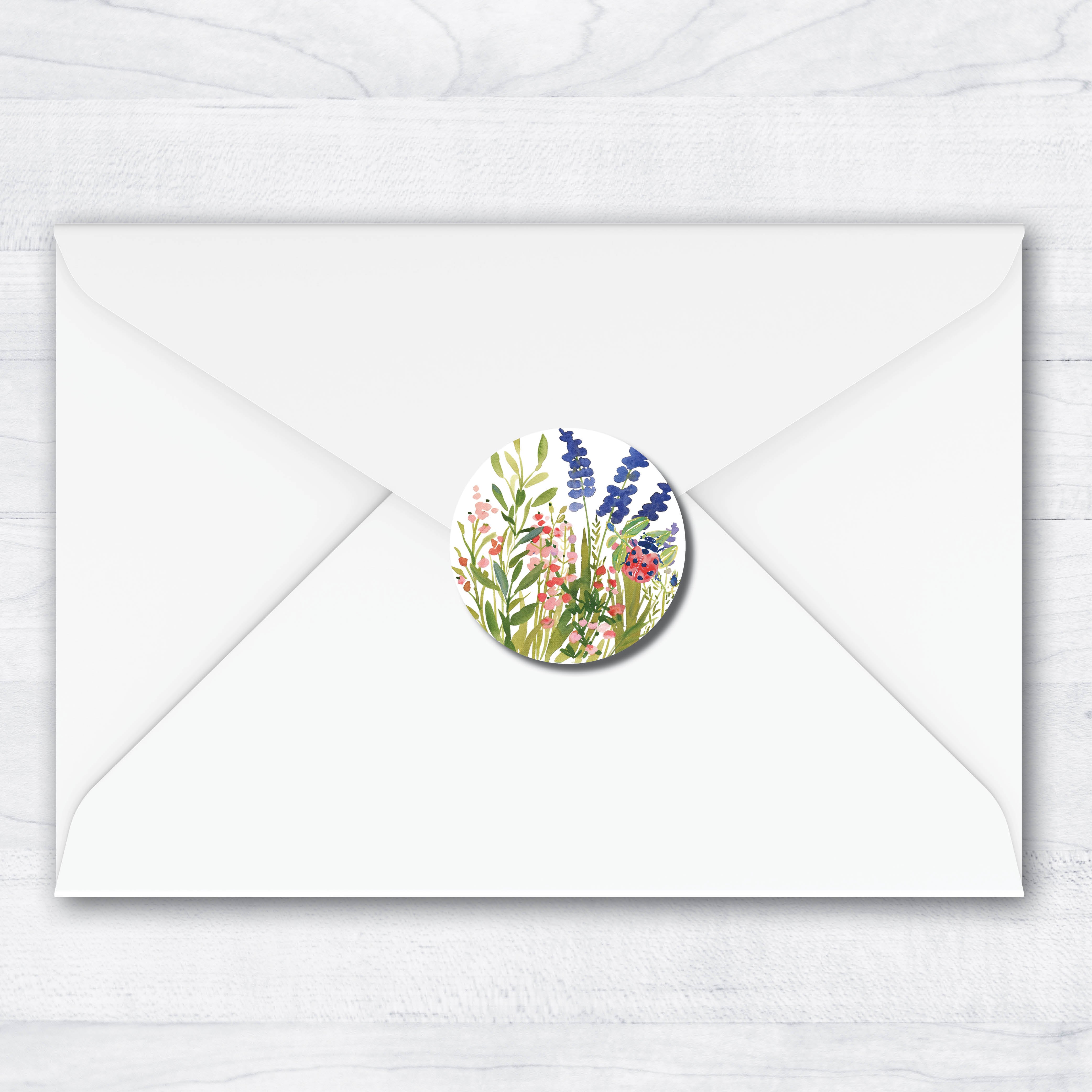 A white envelope centered on a light wood background, with a round floral sticker or seal applied to the front flap. The seal features a watercolor-style botanical design with green stems and leaves, pink flowers, and blue-purple blossoms, and a red ladybug.