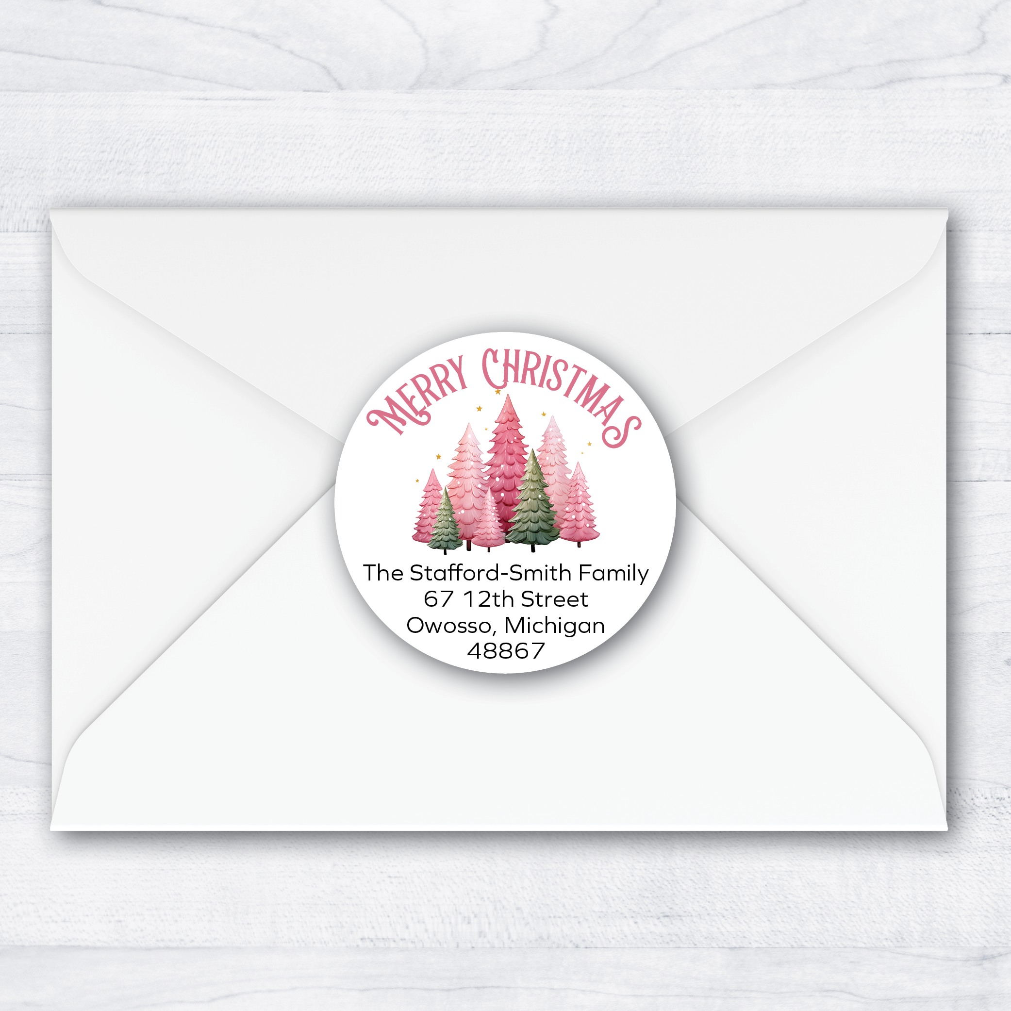 A round Christmas-themed address label with pink and green tree illustrations and the text 'Merry Christmas' and a sample address, shown centered on a white envelope over a light wood background.