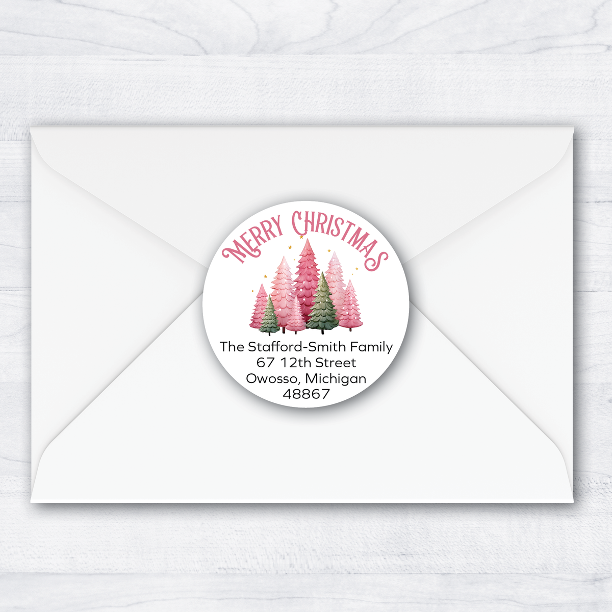 A round Christmas-themed address label with pink and green tree illustrations and the text 'Merry Christmas' and a sample address, shown centered on a white envelope over a light wood background.